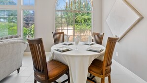 Un coin repas avec une table ronde dressée pour quatre personnes, entourée de chaises en bois, d’un lustre et d’une grande fenêtre en arc offrant une vue sur la verdure.