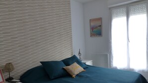 Un dormitorio con cama, mesita de noche, lámpara, un cuadro y ventana con cortinas.