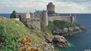 Un castillo de piedra histórico con múltiples torres y muros, ubicado en una costa rocosa con aguas azules y cristalinas.