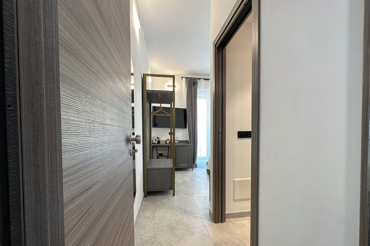 A modern hotel corridor with a wooden door, a bathroom with a mirror, and a seating area with a table and chairs.