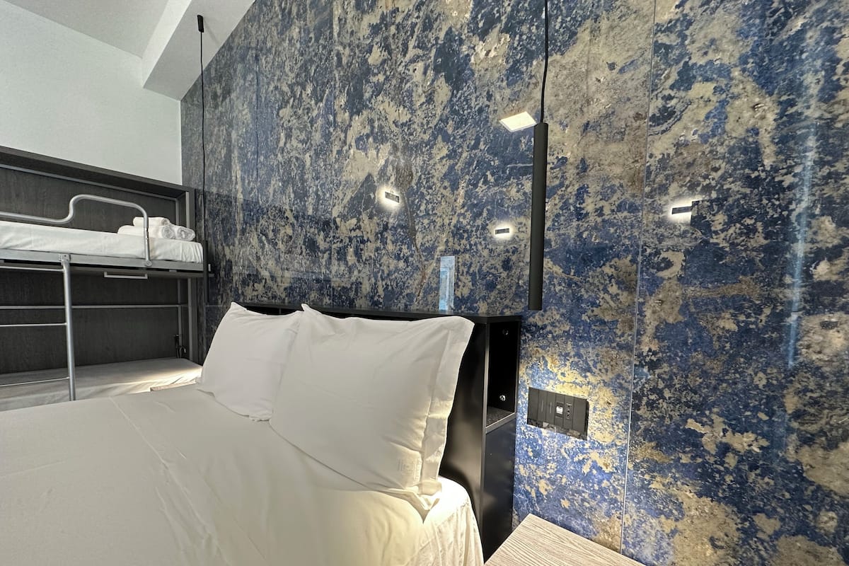A modern hotel room with a bunk bed, a large bed with white linens, and a marble-patterned wall.