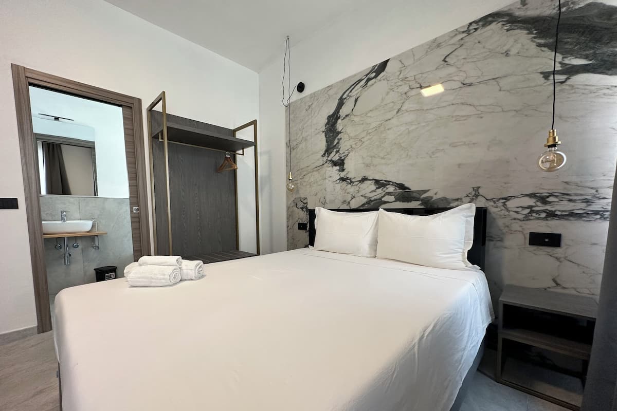A modern hotel room with a large bed, a marble wall, a wooden wardrobe, a bathroom with a sink, and a mirror.