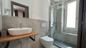 A modern bathroom with a large white sink, a handheld hair dryer, a glass-enclosed shower, and a toilet. - Dolmen Guest House (Alghero)