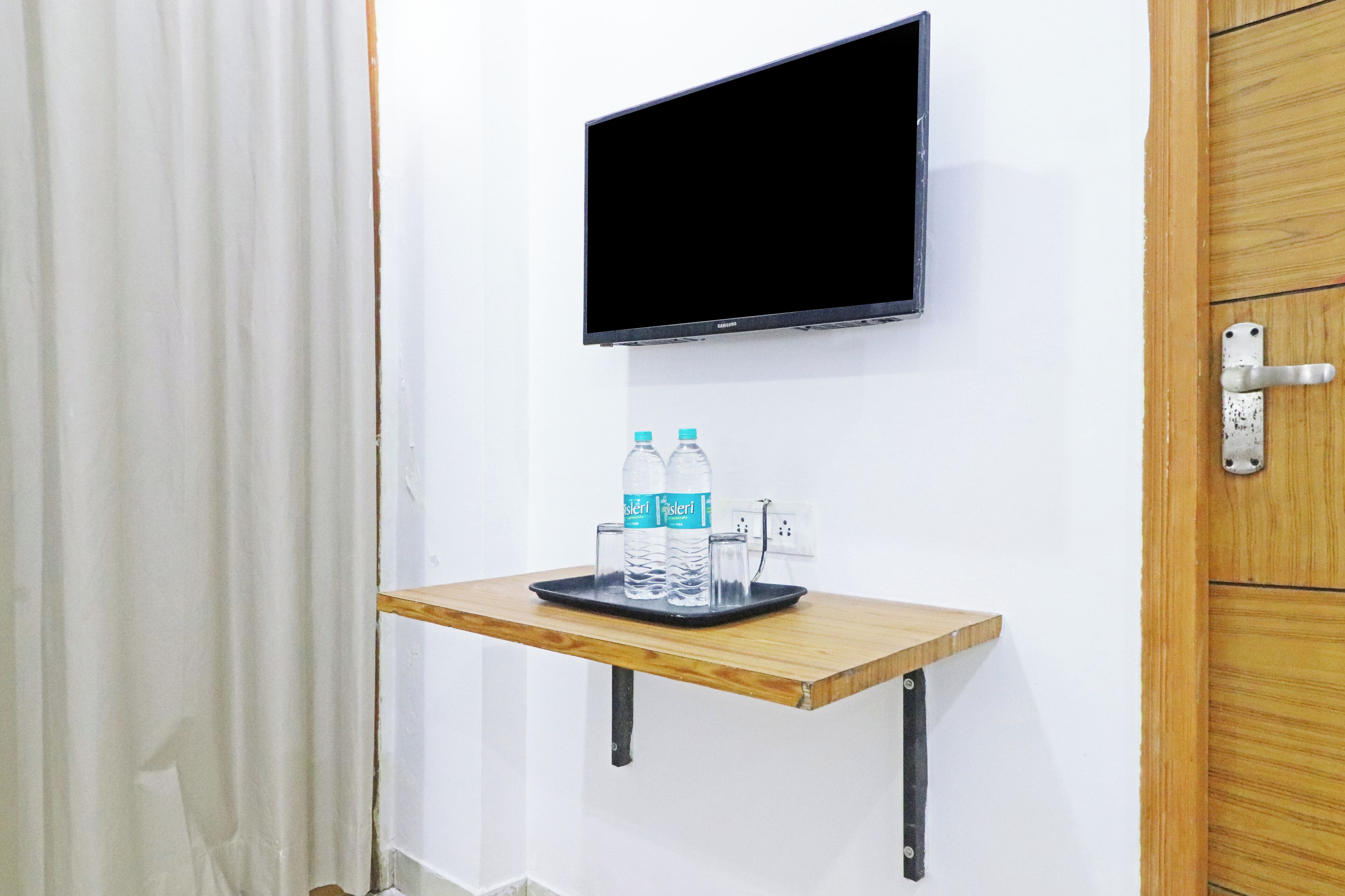 A room with a mounted TV, a wooden shelf with water bottles and glasses, and a wooden door.