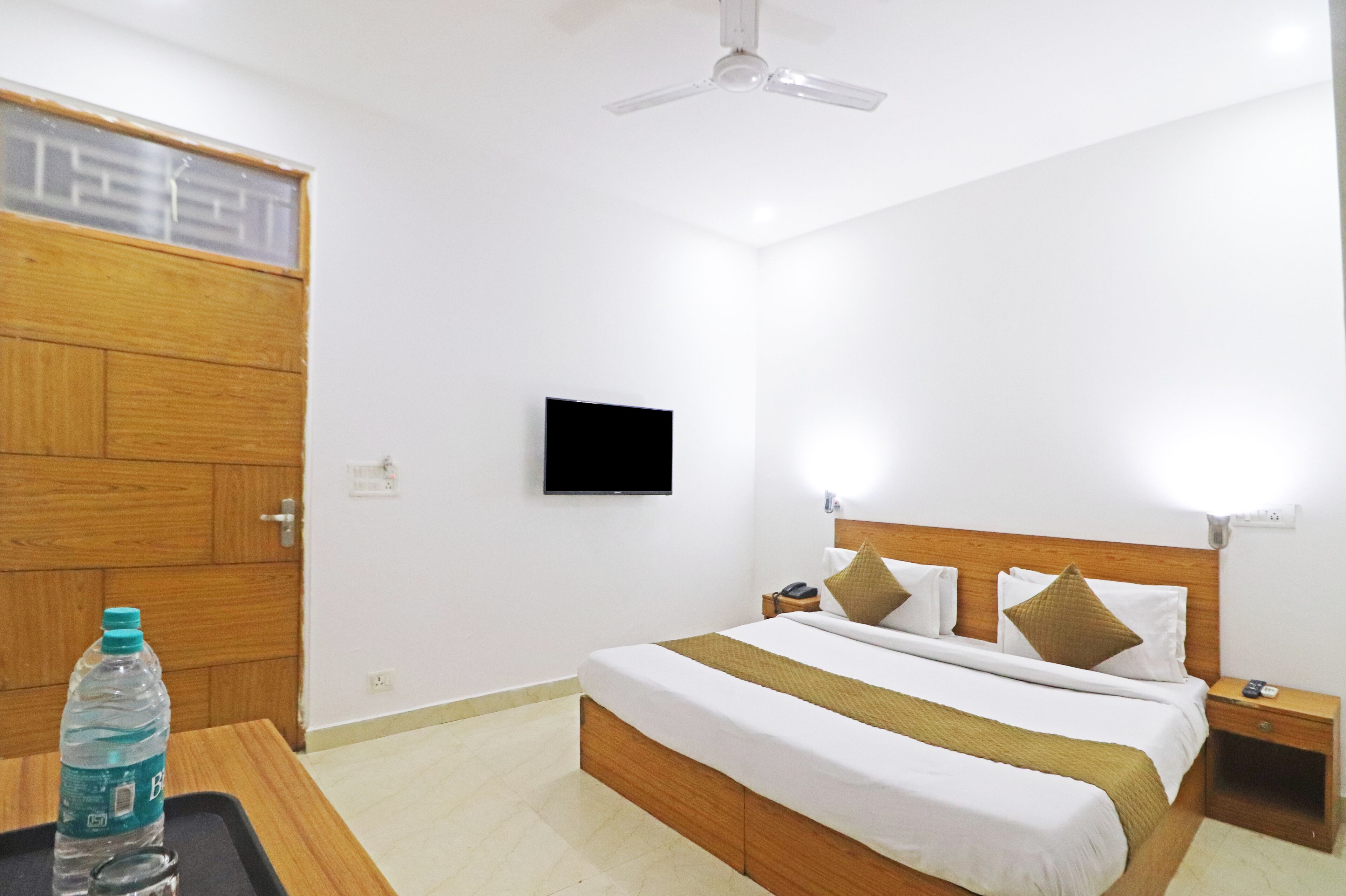 A hotel room with a wooden bed, a flat-screen TV, a bedside table, and a water bottle on a tray.