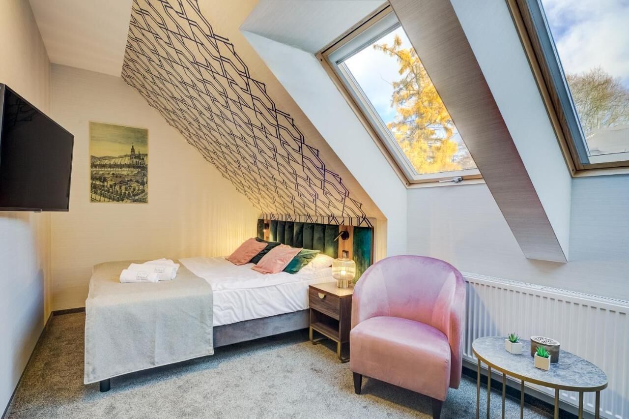 Een slaapkamer met een schuine plafond, een bed, een stoel, een nachtkastje, een lamp en een tafeltje met planten.