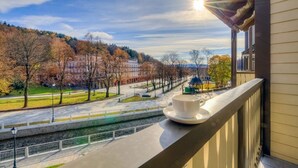 Un balcón con una taza sobre un platillo, con vista a un parque con árboles y un edificio al fondo.
