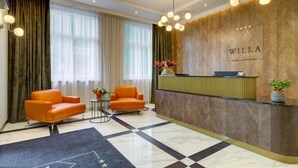 Un área de recepción de hotel con diseño moderno, que incluye un mostrador de recepción, dos sillones naranjas, una mesita auxiliar con un florero y un ventanal con cortinas de tul.