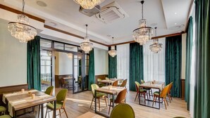 Un comedor de hotel con múltiples mesas y sillas, amplias ventanas con cortinas verdes y candelabros de cristal colgantes.
