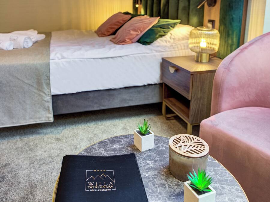 A hotel room with a neatly made bed, a small bedside table with a lamp, a chair, and a decorative pillow.