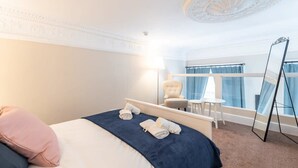 Ett hotellrum med en väl bäddad säng, en stol, ett litet bord, en spegel och utsikt över omgivningarna genom stora fönster.