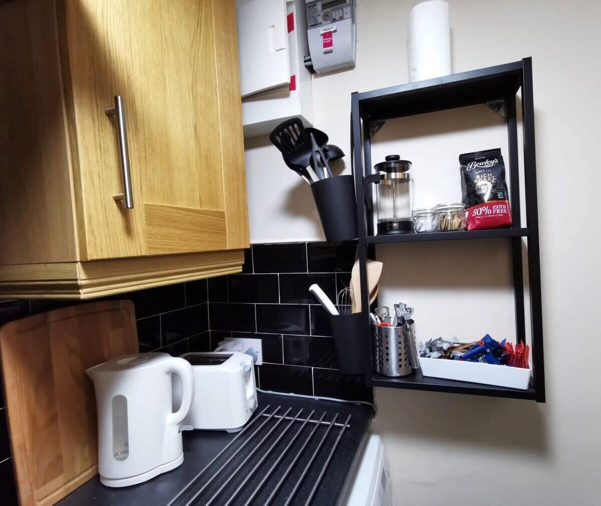 A kitchen with wooden cabinets, a black tiled backsplash, a coffee maker, a kettle, and a shelf with kitchen utensils and a coffee bag.