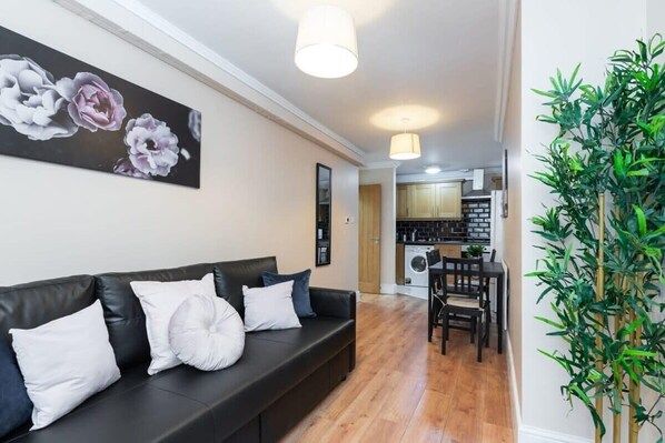 A modern living room with a black leather sofa, a dining table with chairs, a kitchen in the background, and a large green plant. - Lovely Spacious Apartment Right in City Centre! (Dublin 1)