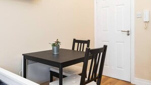 Un pequeño comedor con una mesa de madera oscura y dos sillas, una planta en maceta sobre la mesa, y una puerta blanca con timbre y portero.