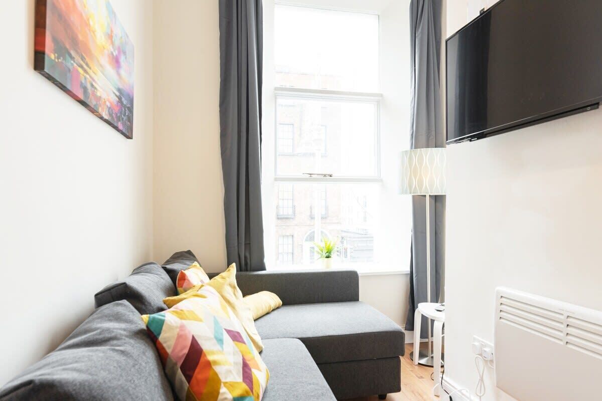 Un salon moderne avec un canapé gris, des coussins colorés, un téléviseur à écran plat, une fenêtre avec des rideaux et un radiateur.