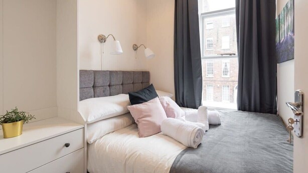 Un lit bien fait, avec une tête de lit grise, du linge de lit blanc et quelques oreillers. On aperçoit une table de chevet sur laquelle sont posés une petite plante et une lampe. Une fenêtre avec des rideaux foncés est visible, et un tableau est accroché au mur.
