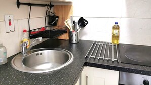 Une cuisine équipée d’un évier en acier inoxydable, d’un plan de travail noir et d’une cuisinière. On y trouve des ustensiles rangés dans un porte-ustensiles et une bouteille de liquide vaisselle posée sur le plan de travail.