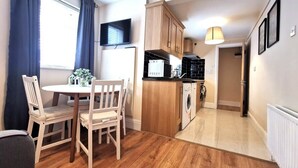 Una cocina compacta con microondas, estufa y lavabo. Hay una mesa de comedor con sillas y una planta pequeña encima. El ambiente tiene piso de madera y una ventana con cortinas.