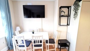 En matplats med ett vitt bord och stolar, en platt-tv monterad på väggen, ett litet svart sidobord och en hylla med dekorativa föremål.