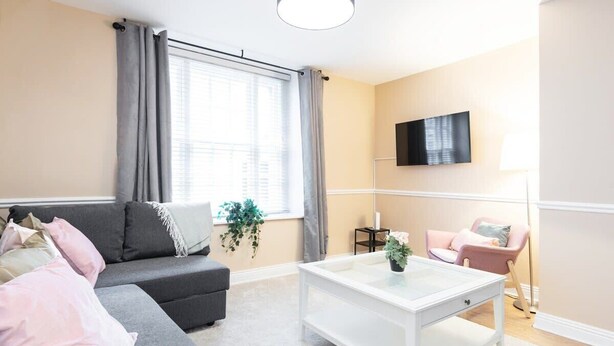 Un soggiorno moderno con un divano grigio, un tavolino da caffè bianco, una poltrona rosa e una TV a parete.