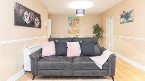 Un soggiorno moderno con un divano grigio, cuscini rosa e neri, e una coperta bianca. Si notano dipinti floreali sulle pareti e un lampadario a soffitto.