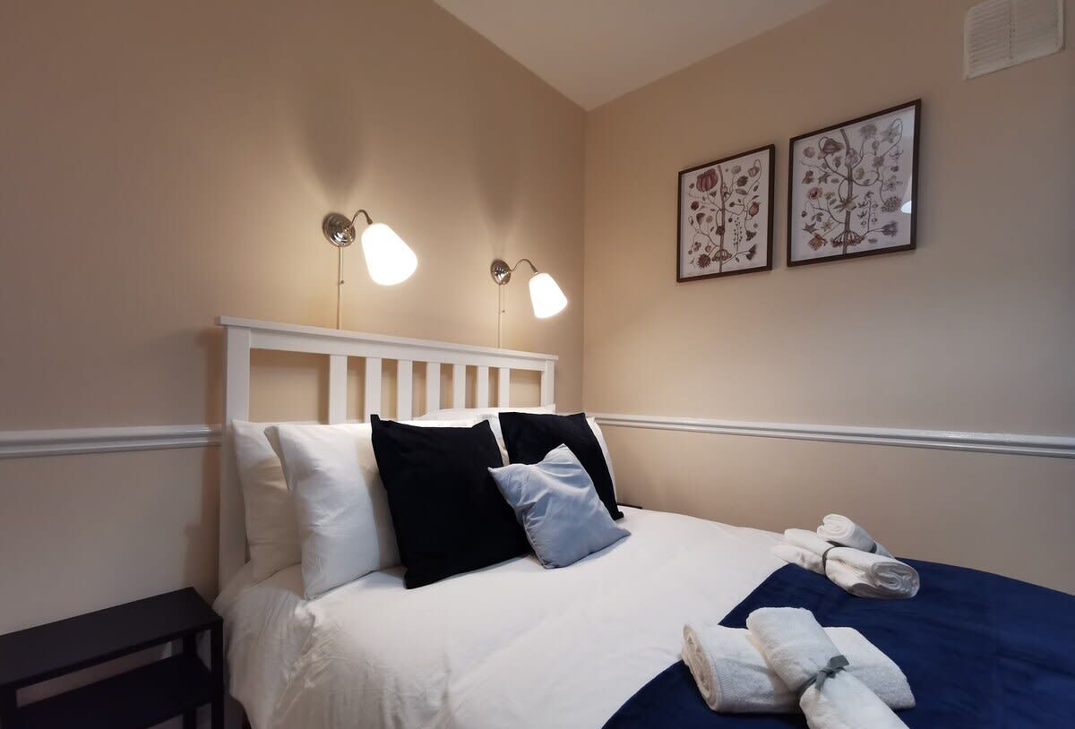 A neatly made bed with white and black pillows, a blue bedspread, and rolled towels on the side. There are two wall-mounted light fixtures and two framed floral artworks above the bed.