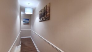 Un couloir étroit doté d’un plancher en bois et de moulures blanches, d’un luminaire au plafond et de deux œuvres d’art encadrées accrochées aux murs.