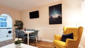 Un salon avec un fauteuil jaune, une table blanche ornée d’un vase de fleurs, un coin repas avec des chaises, un téléviseur fixé au mur et un tableau représentant un ciel nocturne.