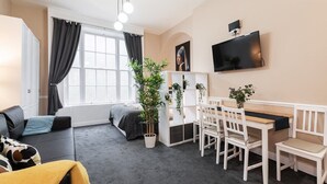 Un salon moderne comprenant un canapé, une table à manger avec des chaises, un téléviseur fixé au mur et une grande fenêtre avec des rideaux.