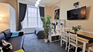 Un salon moderne comprenant un canapé, une table à manger avec des chaises, un téléviseur fixé au mur et une grande fenêtre avec des rideaux.
