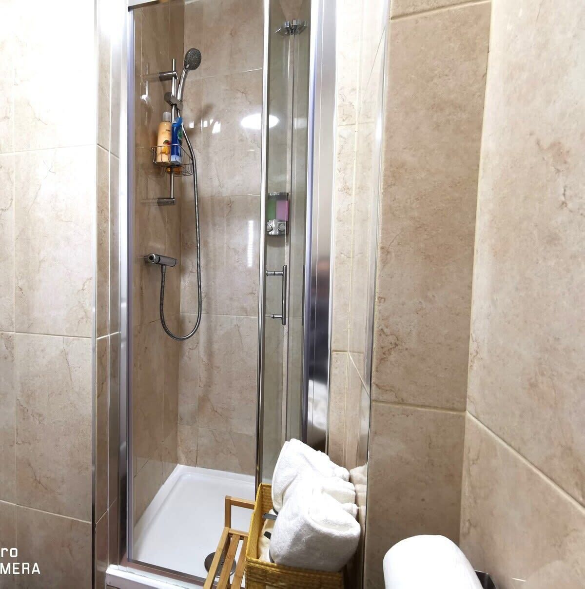 Zona de ducha con mampara de vidrio, cabezal de ducha manual, grifo empotrado en la pared y toallero con una toalla doblada.