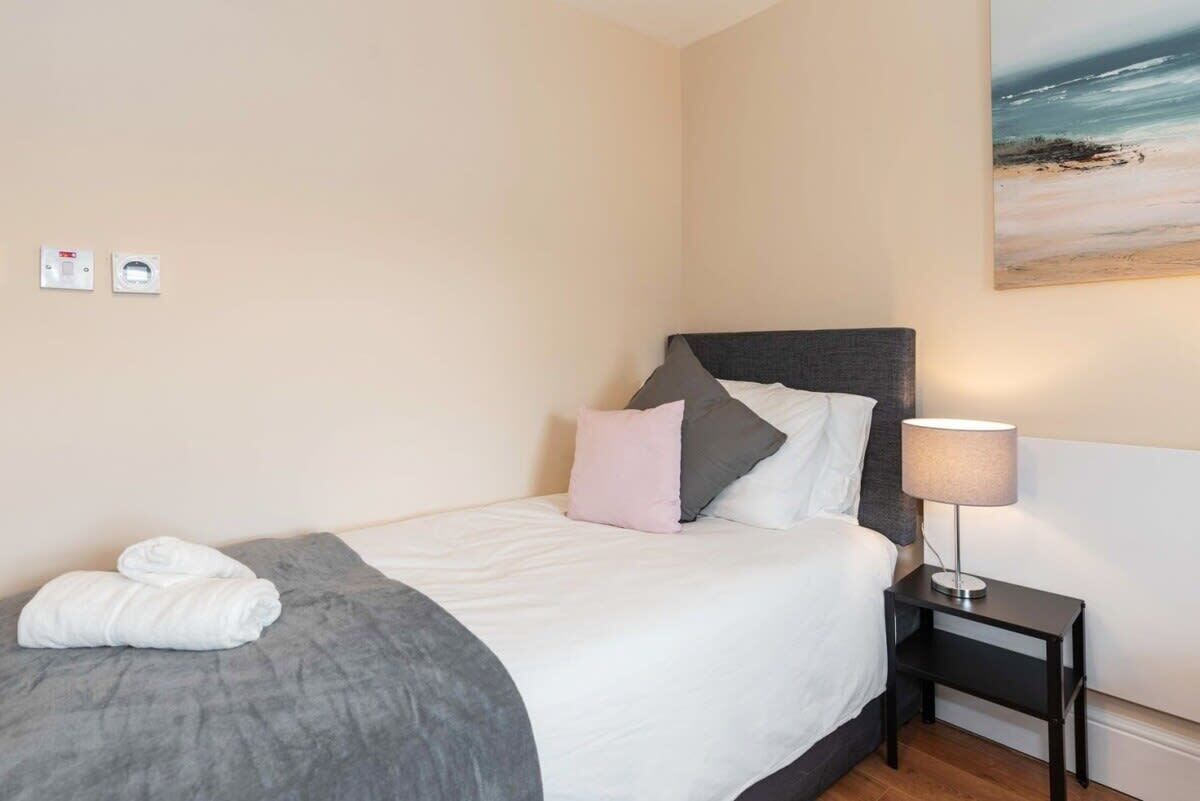 A neatly made bed with white and grey bedding, a grey throw blanket, and a pair of white towels. There is a bedside lamp on a small table beside the bed.