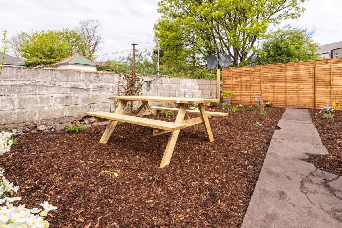 A wooden picnic table in a garden area with mulch, a concrete path, and a wooden fence.