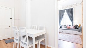 Een eetgedeelte met een witte tafel en stoelen, een bank met kussens, en een raam met gordijnen.