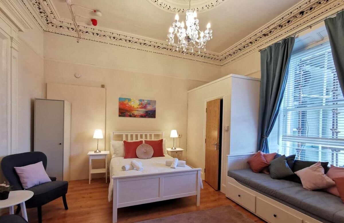A bedroom with a bed, bedside tables, a chair, a sofa, a chandelier, and a painting on the wall.