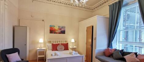 A bedroom with a bed, bedside tables, a chair, a sofa, a chandelier, and a painting on the wall.