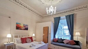 Een slaapkamer met een bed, nachtkastjes, een bank met kussens, een kroonluchter en een schilderij aan de muur.