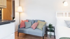 Een moderne, compacte leefruimte met een grijze bank, een klein nachtkastje met een lamp en een wit bed.