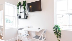 Een lichte kamer met een wit bureau, twee witte stoelen, een wandtelevisie en een raam met uitzicht.