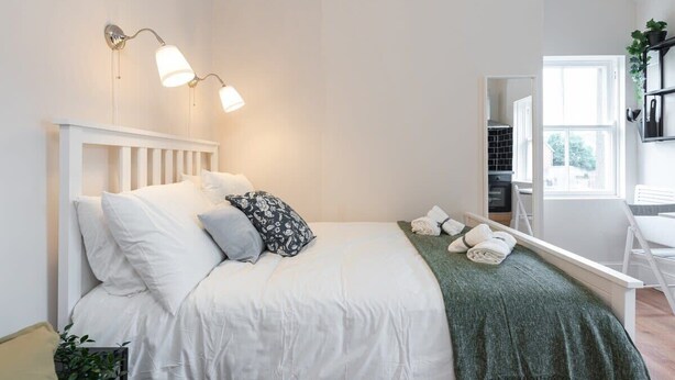 Een netjes opgemaakte slaapkamer met een wit bed, een groene plaid en een nachtkastje met handdoeken.