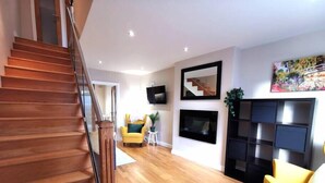 A modern living room with a wooden staircase, a flat-screen TV, a fireplace, and a yellow armchair.