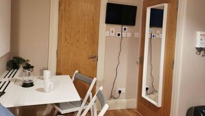 Ein kleines Zimmer mit einer Holztür, einem weißen Tisch mit zwei Stühlen, einem an der Wand befestigten Fernseher und einer Steckdose mit Stecker.