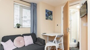 Quarto pequeno e moderno com sofá cinza, mesa branca e área de chuveiro.