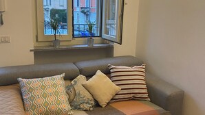A cozy living room with a grey sofa, patterned cushions, a small potted plant, and a view of buildings through a window. - Navigli bilocale  (Milan)
