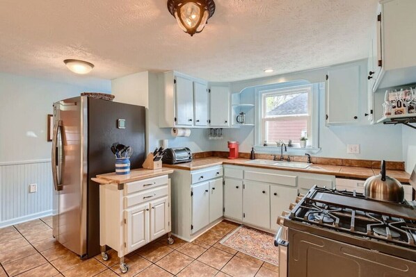 Compact kitchen with a gas stove, refrigerator, and toaster, featuring a window above the sink and a tiled floor.