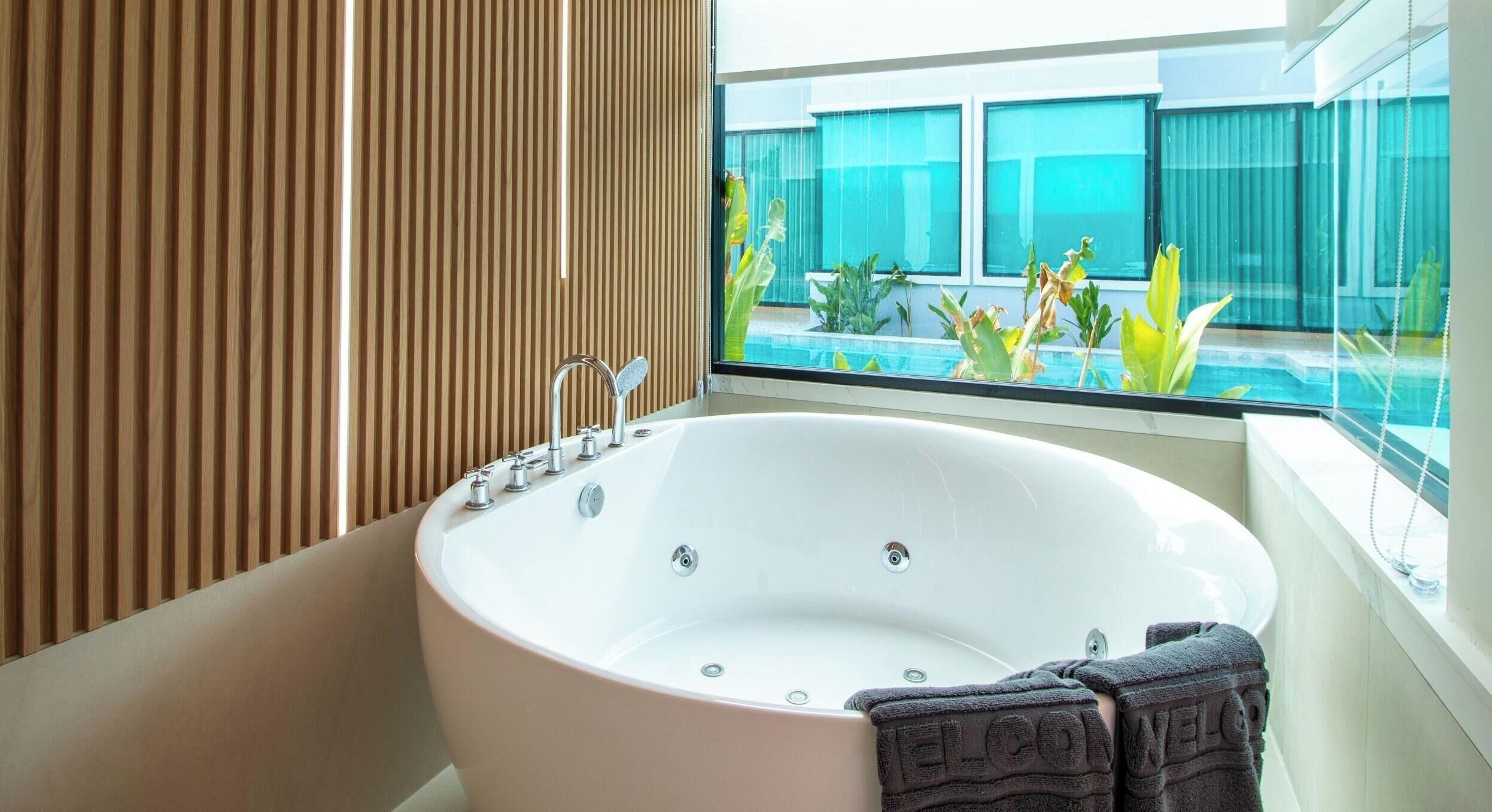 A modern bathroom with a freestanding bathtub, a window with a view of a pool and greenery, and a towel rack with towels.