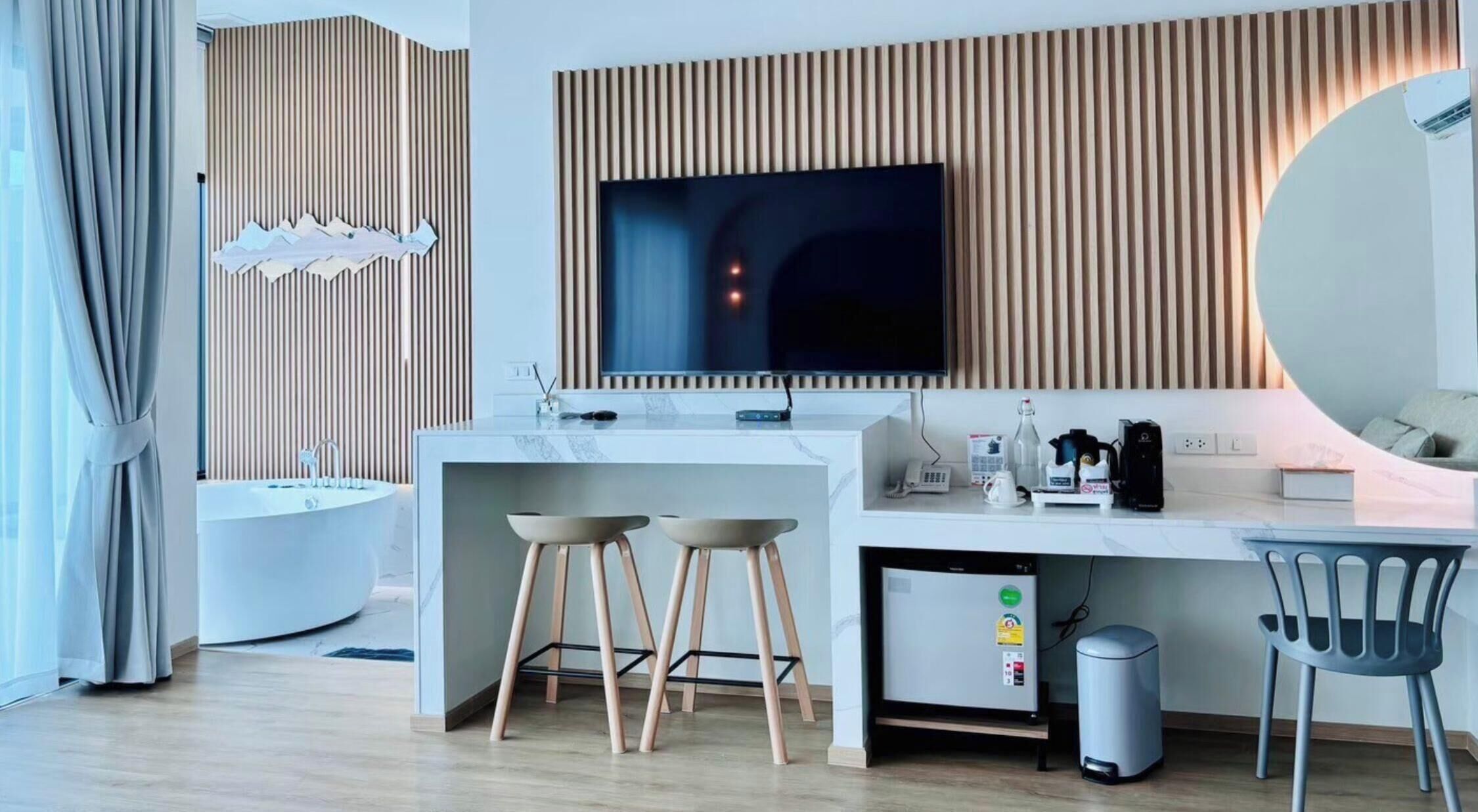 A modern hotel room with a central island, a large flat-screen TV, a round mirror, a desk with a coffee maker, and a trash bin.