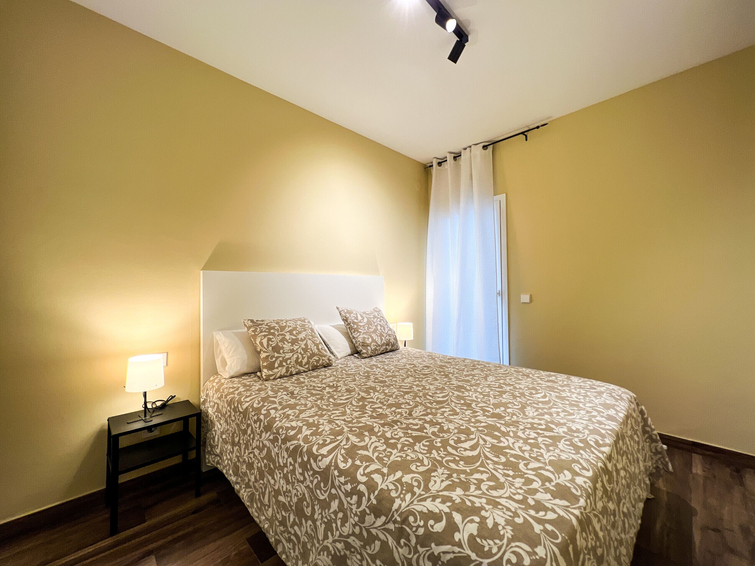 A bedroom with a large bed, a nightstand, a lamp, and a window with curtains.
