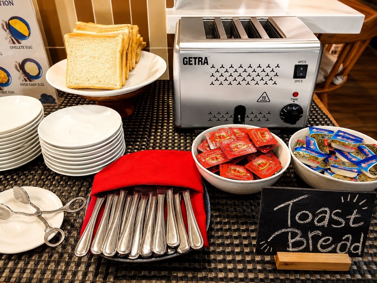 A breakfast buffet setup with a toaster, plates, cutlery, and bread.