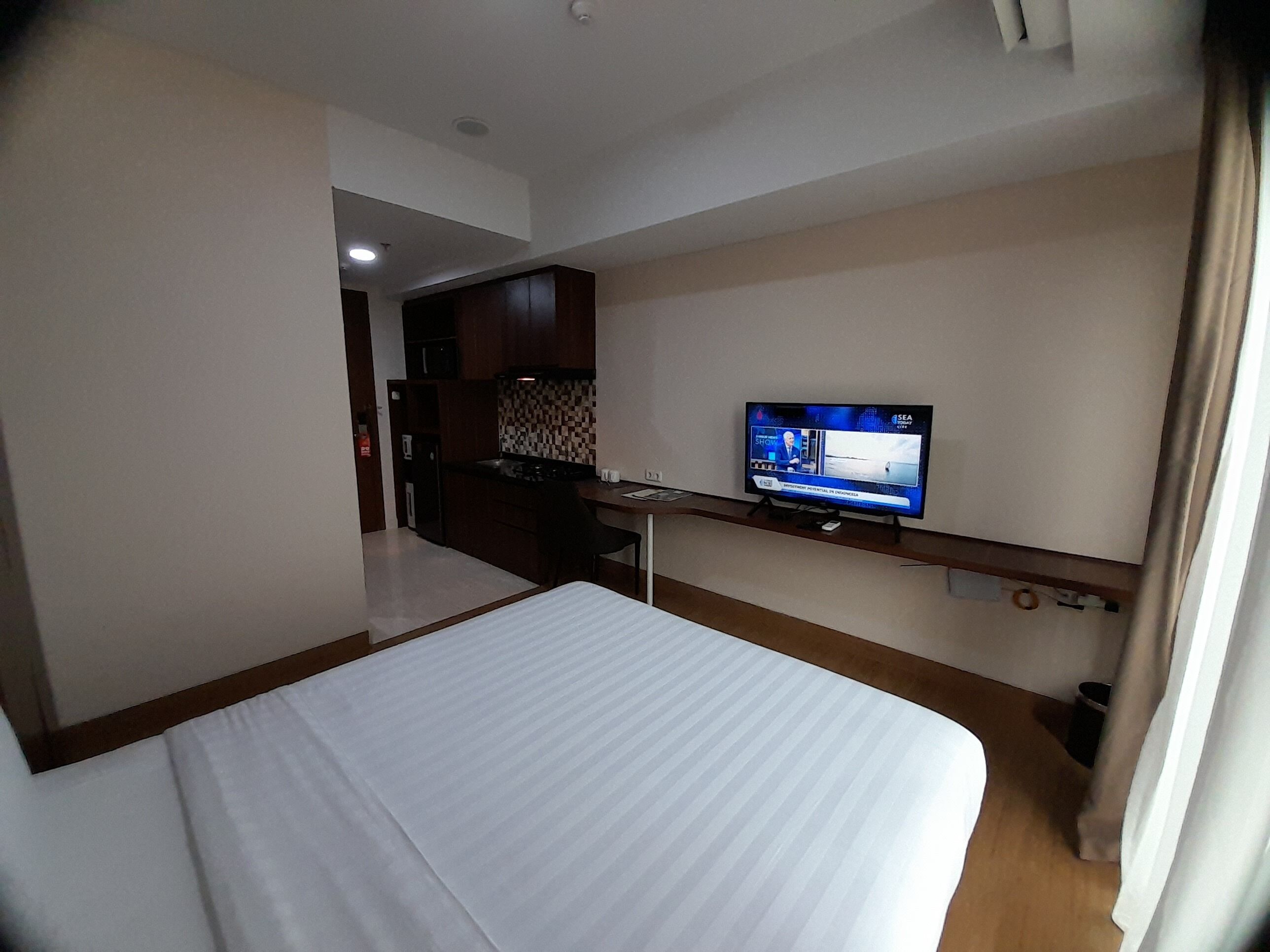 A hotel room with a bed, a television on a stand, a kitchenette, and a bathroom visible through an open door.